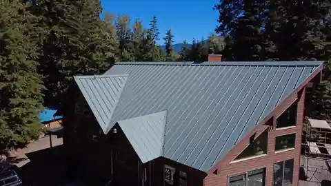 Grey standing-seam metal roof on a log A-frame among pines, seen by drone