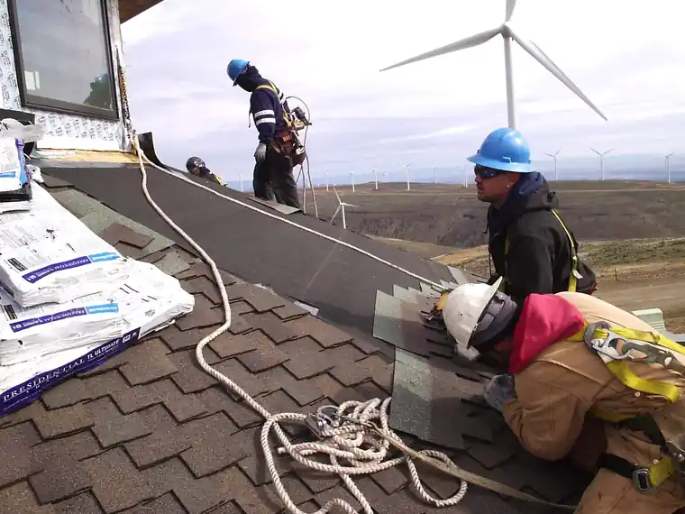 Crew in hard hats shingling a roof with wind turbines behind